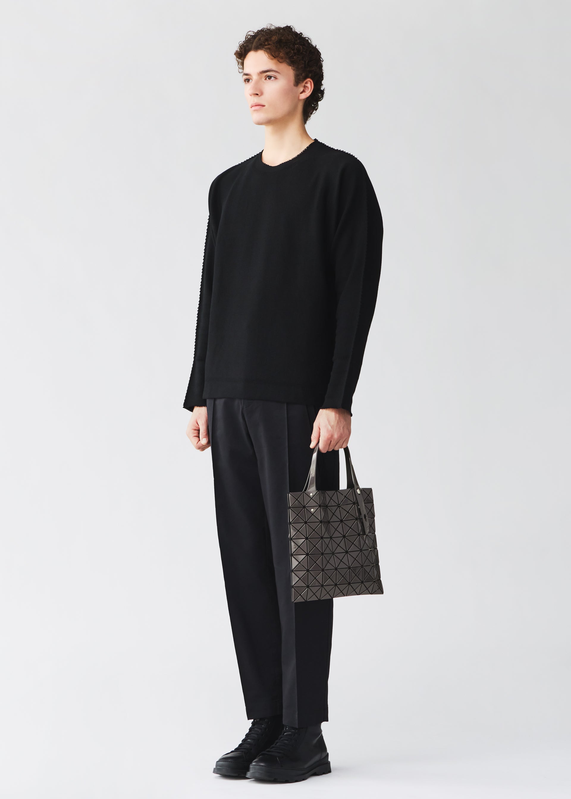Zoomed view A person stands against a plain light background, dressed in a black sweater, black pants, and black shoes, holding a BAO BAO ISSEY MIYAKE PRISM GLOSS tote bag with a geometric pattern and an enamel-like finish in their left hand.