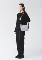 A person dressed in black stands facing left against a plain light background, holding the MUSETTE cross-body bag by BAO BAO ISSEY MIYAKE—a white bag featuring a geometric pattern and crafted from a leather-like material.