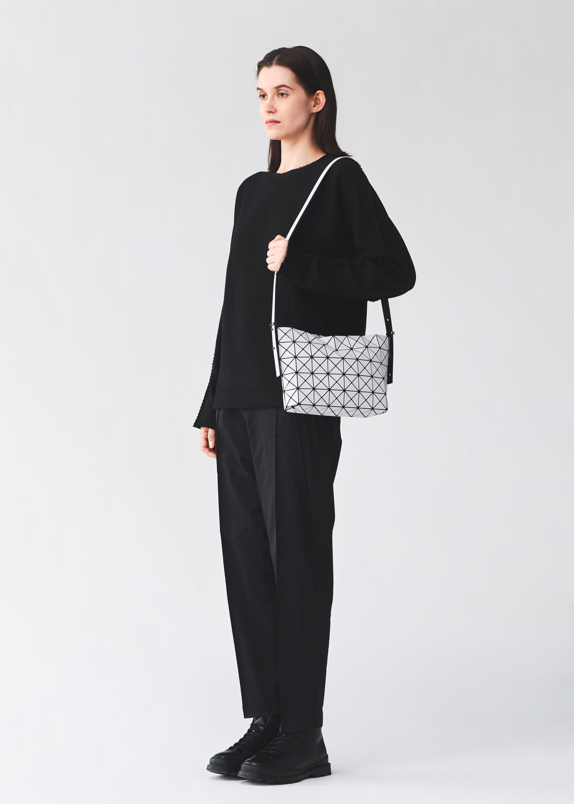 Zoomed view A person dressed in black stands facing left against a plain light background, holding the MUSETTE cross-body bag by BAO BAO ISSEY MIYAKE—a white bag featuring a geometric pattern and crafted from a leather-like material.