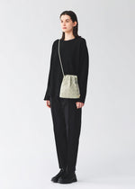 A person stands against a plain background, dressed in a loose black sweater, black pants, and black shoes, carrying the BAO BAO ISSEY MIYAKE WRING CLAY—a small nubuck-like geometric-patterned shoulder bag.