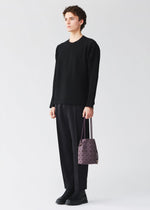 A person with short curly hair wears a black sweater, black pants, and black shoes, holding the BAO BAO ISSEY MIYAKE WRING CLAY drawstring bag in a purple geometric pattern with white handles, standing against a plain light background.