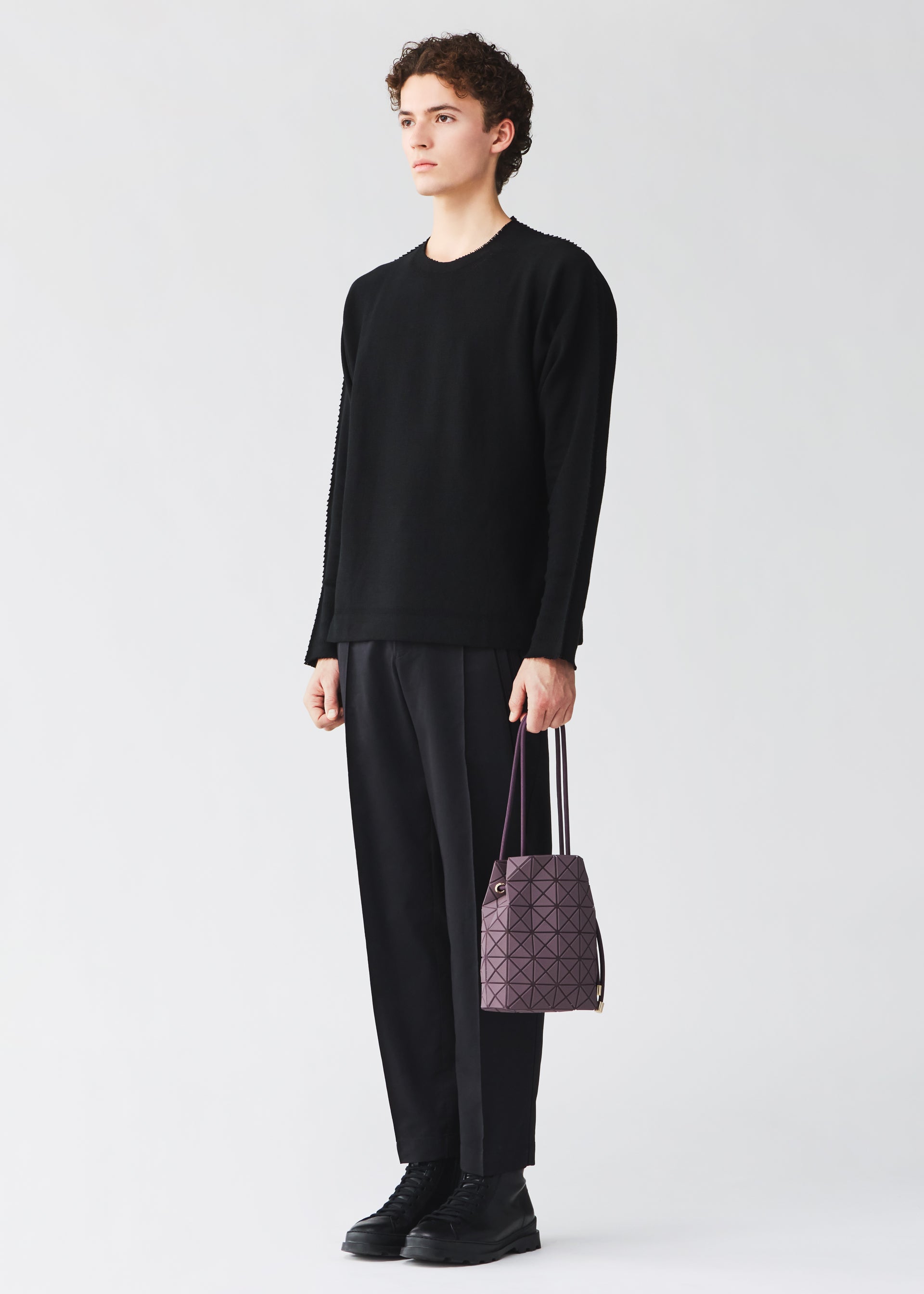 Zoomed view A person with short curly hair wears a black sweater, black pants, and black shoes, holding the BAO BAO ISSEY MIYAKE WRING CLAY drawstring bag in a purple geometric pattern with white handles, standing against a plain light background.