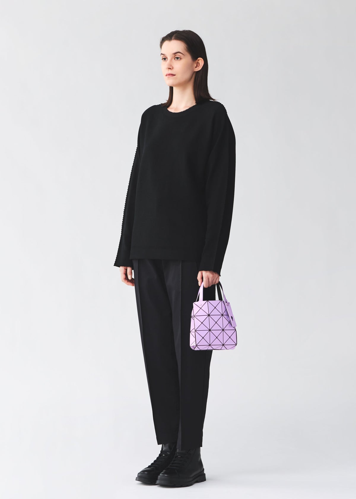 A person stands against a plain background wearing a black sweater, black pants, and black boots, holding the BAO BAO ISSEY MIYAKE CARAT handbag—a small lavender bag with a geometric pattern and an inner pocket—in their left hand. view 9 of 9