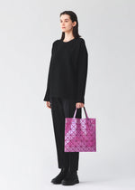 A person with long dark hair, dressed in black, stands against a plain light background holding a glossy pink LUCENT ONE-TONE geometric tote bag by BAO BAO ISSEY MIYAKE.