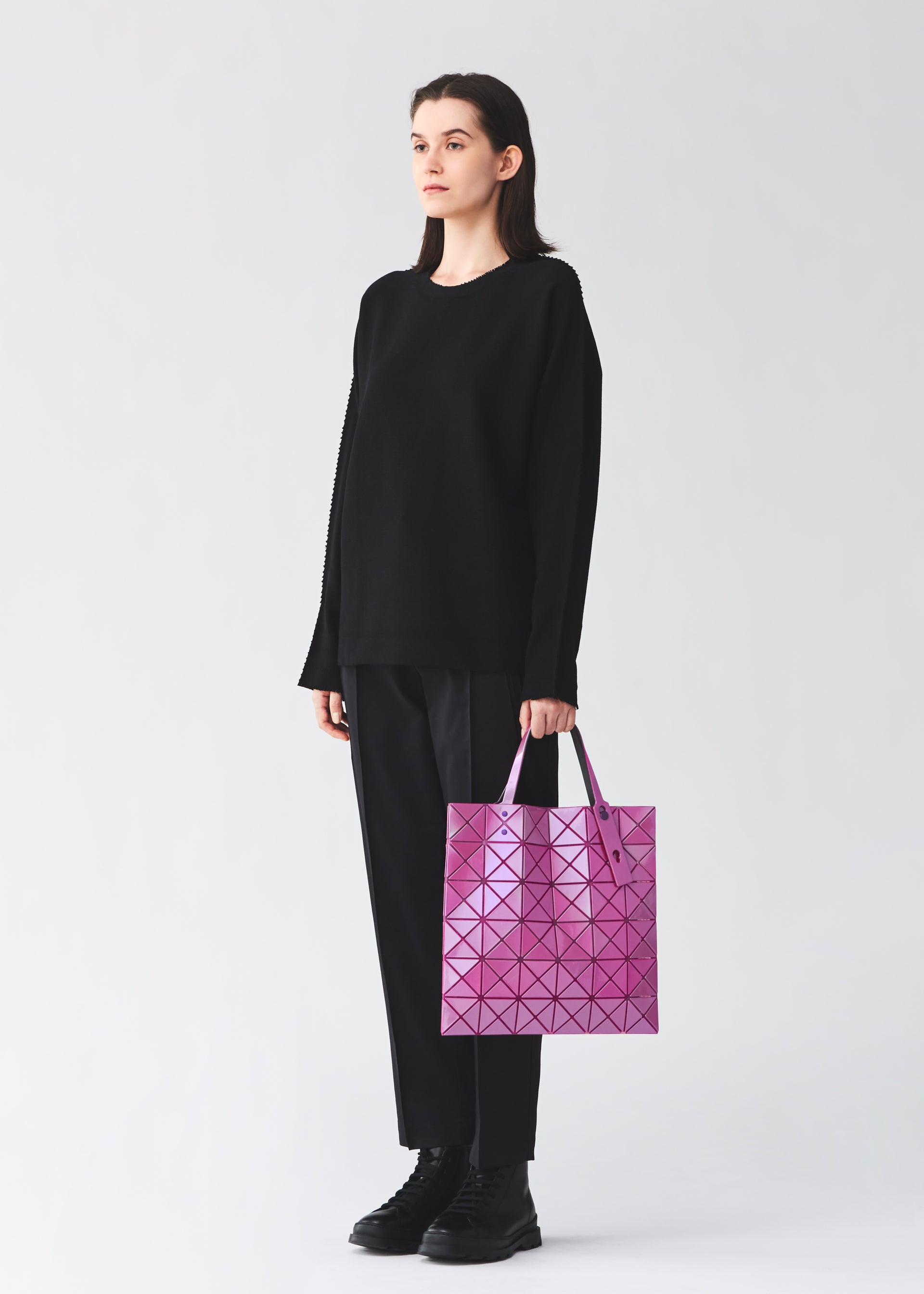 Zoomed view A person with long dark hair, dressed in black, stands against a plain light background holding a glossy pink LUCENT ONE-TONE geometric tote bag by BAO BAO ISSEY MIYAKE.