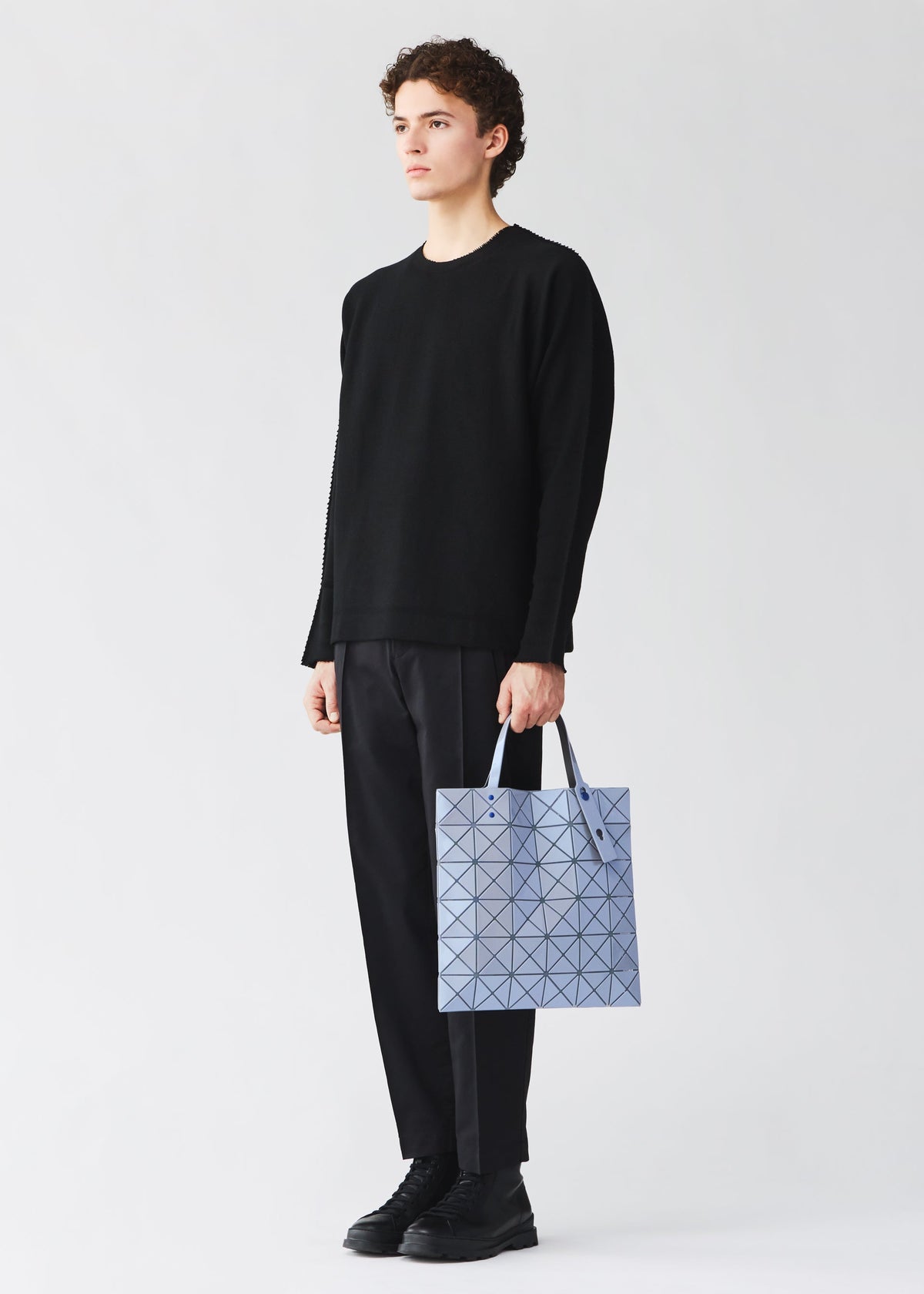 A person in a loose black sweater, black pants, and black shoes holds a light blue LUCENT ONE-TONE pouch by BAO BAO ISSEY MIYAKE against a plain white background. view 10 of 10