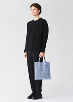 A person in a loose black sweater, black pants, and black shoes holds a light blue LUCENT ONE-TONE pouch by BAO BAO ISSEY MIYAKE against a plain white background.