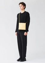 A person with short curly hair, in a black long-sleeved top, loose black pants, and black shoes, stands against a plain light background wearing the yellow LUCENT ONE-TONE tote bag by BAO BAO ISSEY MIYAKE crossbody.