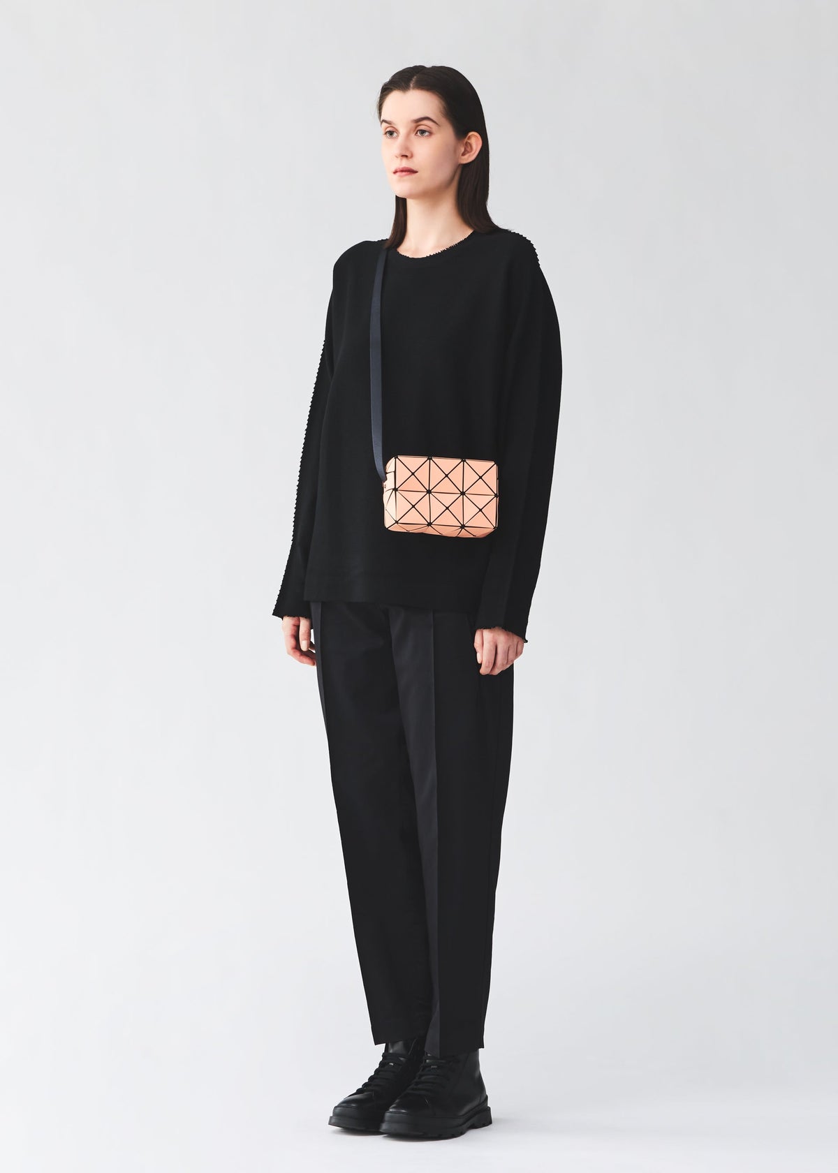 A person stands against a light background, wearing a loose black sweater, black pants, black shoes, and carries the BAO BAO ISSEY MIYAKE CUBOID—a small minimalist crossbody bag featuring a geometric pattern and an adjustable strap. view 7 of 8