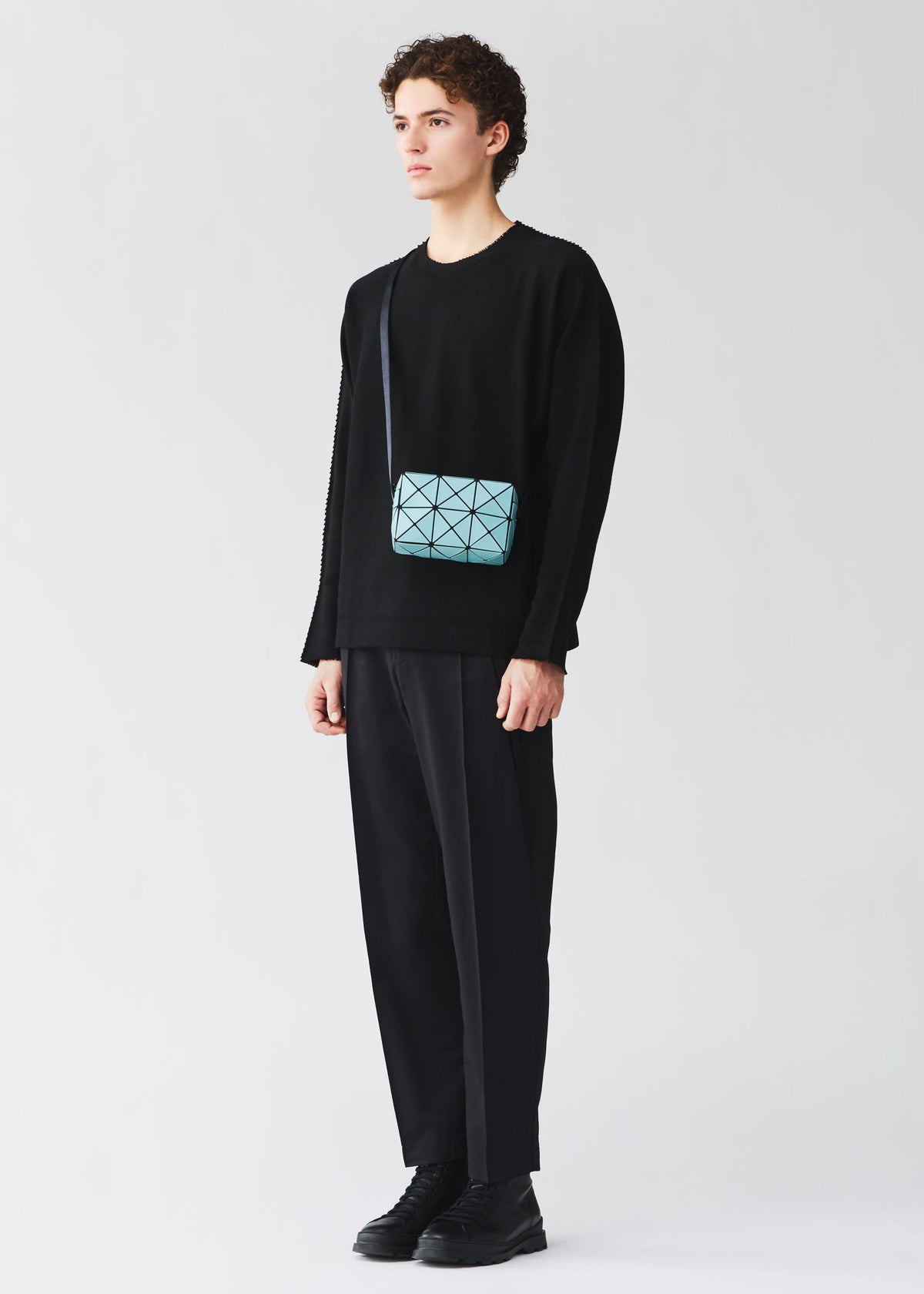 Against a plain background, a person wears a black sweater, black pants, and black shoes. They complete the look with the BAO BAO ISSEY MIYAKE CUBOID bag—a small, light blue minimalist piece featuring an adjustable strap worn crossbody. view 8 of 8