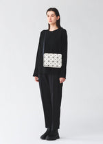 A person stands against a plain background wearing a loose black outfit and black shoes, carrying the BAO BAO ISSEY MIYAKE CUBOID shoulder bag—a geometric white bag with a black strap featuring a minimalist design and cuboid shape.
