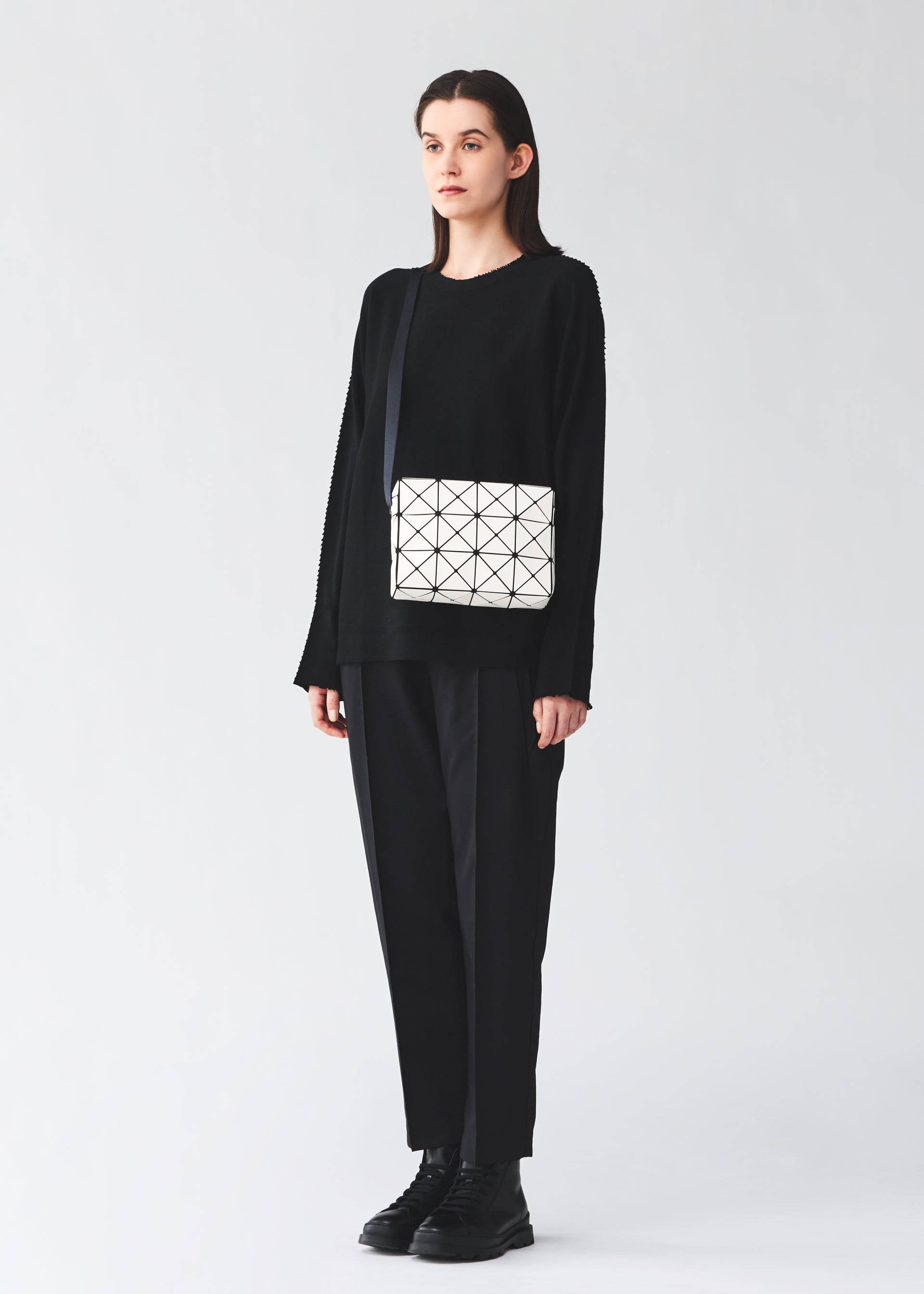 Zoomed view A person stands against a plain background wearing a loose black outfit and black shoes, carrying the BAO BAO ISSEY MIYAKE CUBOID shoulder bag—a geometric white bag with a black strap featuring a minimalist design and cuboid shape.