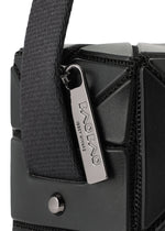 Close-up of the CUBOID bag by BAO BAO ISSEY MIYAKE, showcasing its black geometric cuboid shape and a metal zipper pull engraved with the brand name, attached to a textured fabric handle. This shoulder bag features a minimalist design that emphasizes its modern aesthetic.