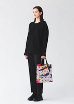 A person in black stands against a plain background, holding the BAO BAO ISSEY MIYAKE WARP tote, featuring a geometric design and colorful iridescent enamel accents.