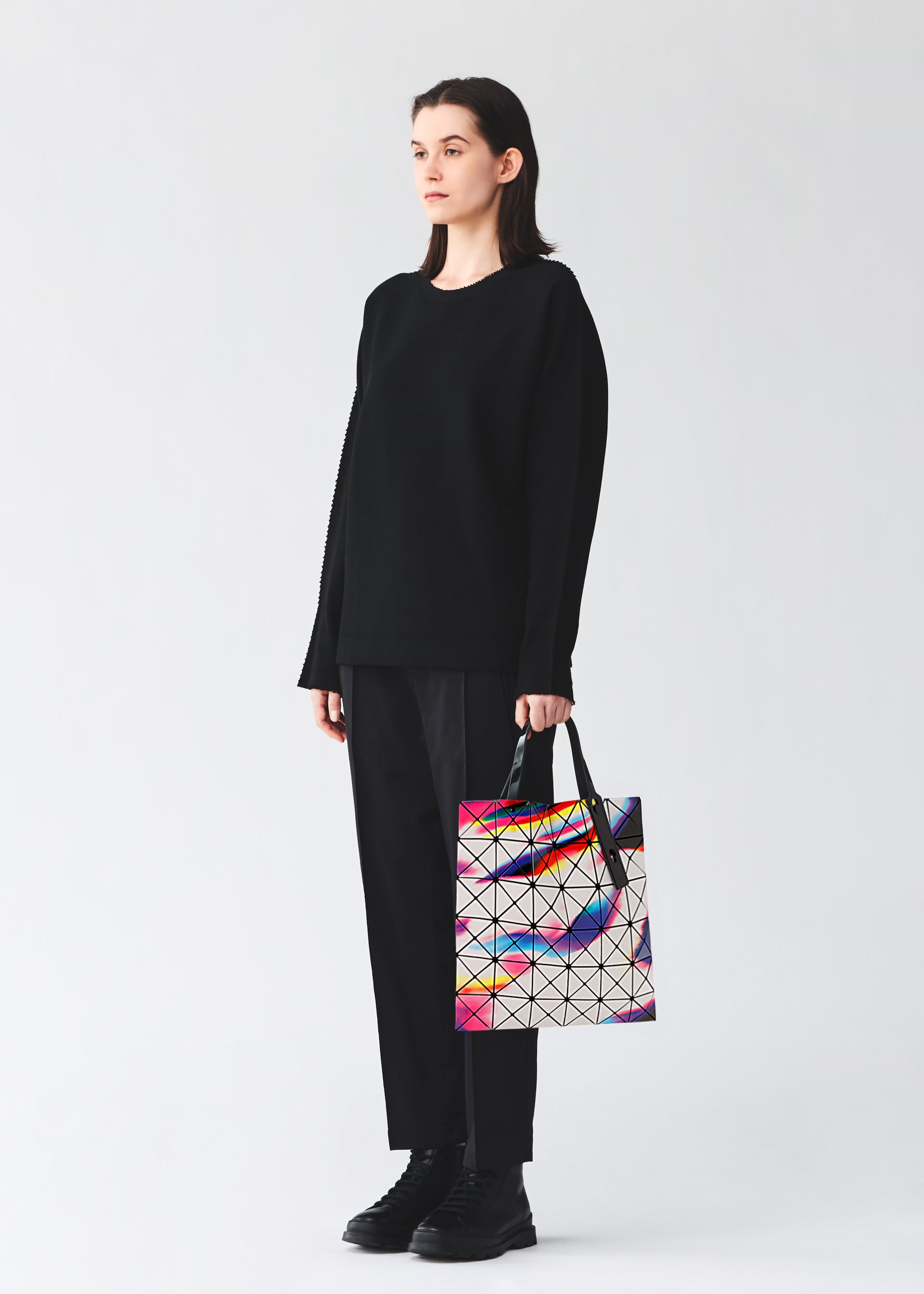 Zoomed view A person in black stands against a plain background, holding the BAO BAO ISSEY MIYAKE WARP tote, featuring a geometric design and colorful iridescent enamel accents.
