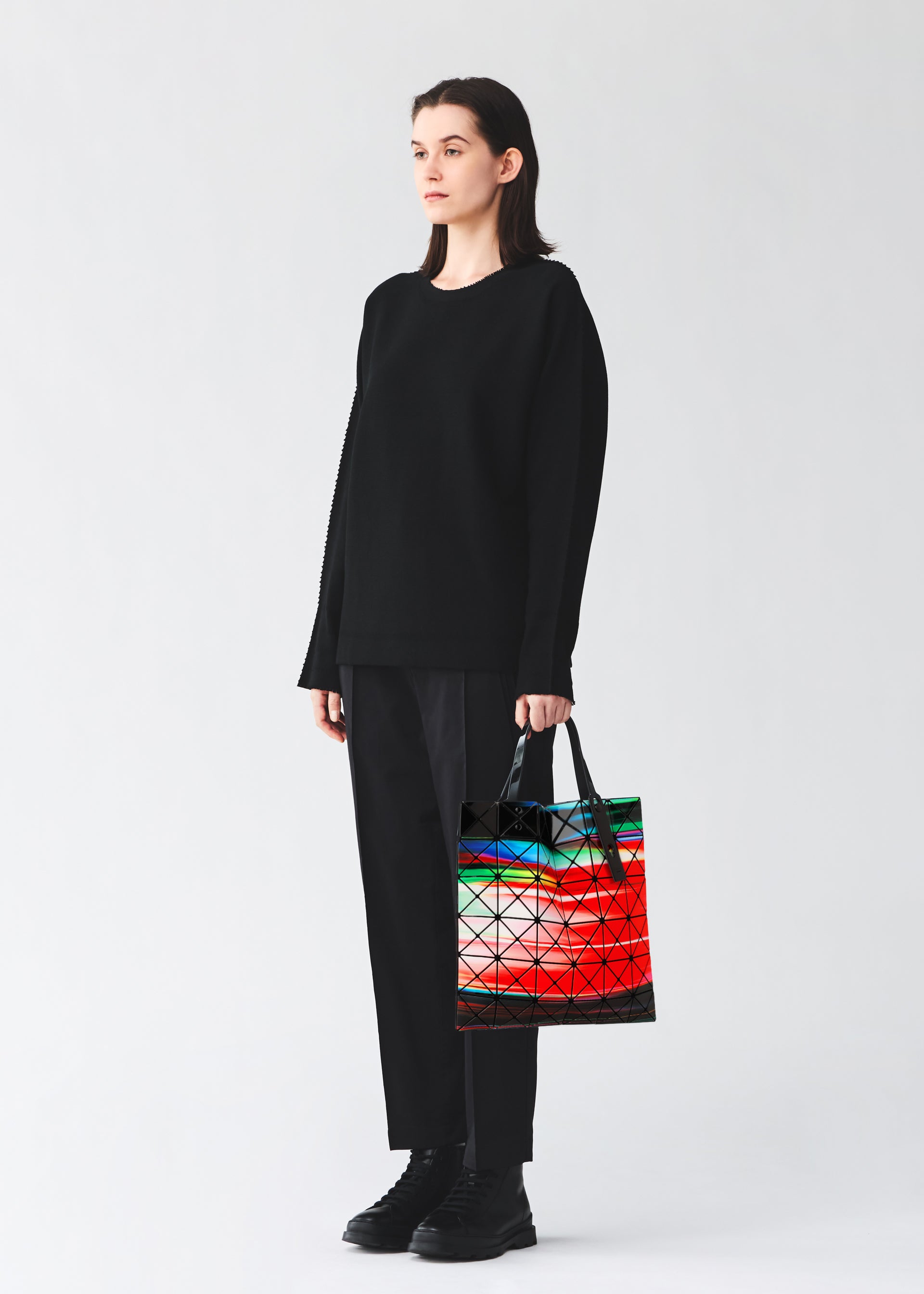 Zoomed view A woman in all black stands against a plain backdrop, holding the BAO BAO ISSEY MIYAKE WARP tote—a geometric bag with striking enamel material and a colorful abstract pattern in red, green, and white.
