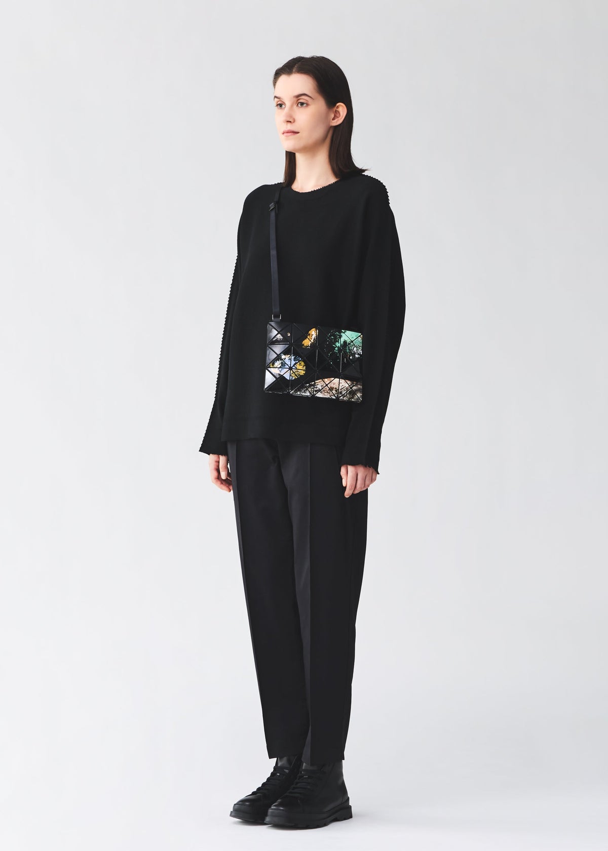 A person in loose black clothing and black boots stands against a plain light background, holding the BAO BAO ISSEY MIYAKE COSMIC shoulder bag, which features an adjustable strap and a colorful forest and bird design. view 9 of 10