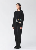 A person in loose black clothing and black boots stands against a plain light background, holding the BAO BAO ISSEY MIYAKE COSMIC shoulder bag, which features an adjustable strap and a colorful forest and bird design.