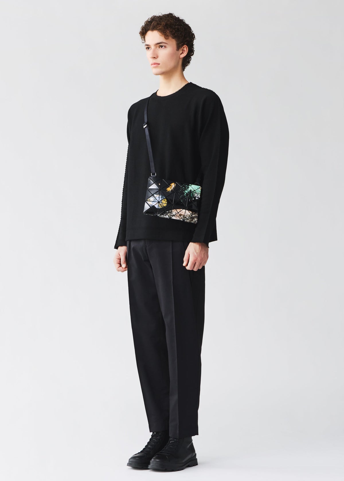 A person stands against a plain background, wearing a black sweater, black pants, black shoes, and carrying the BAO BAO ISSEY MIYAKE COSMIC shoulder bag, which features a geometric design with colorful accents and an adjustable strap for added comfort and versatility. view 10 of 10