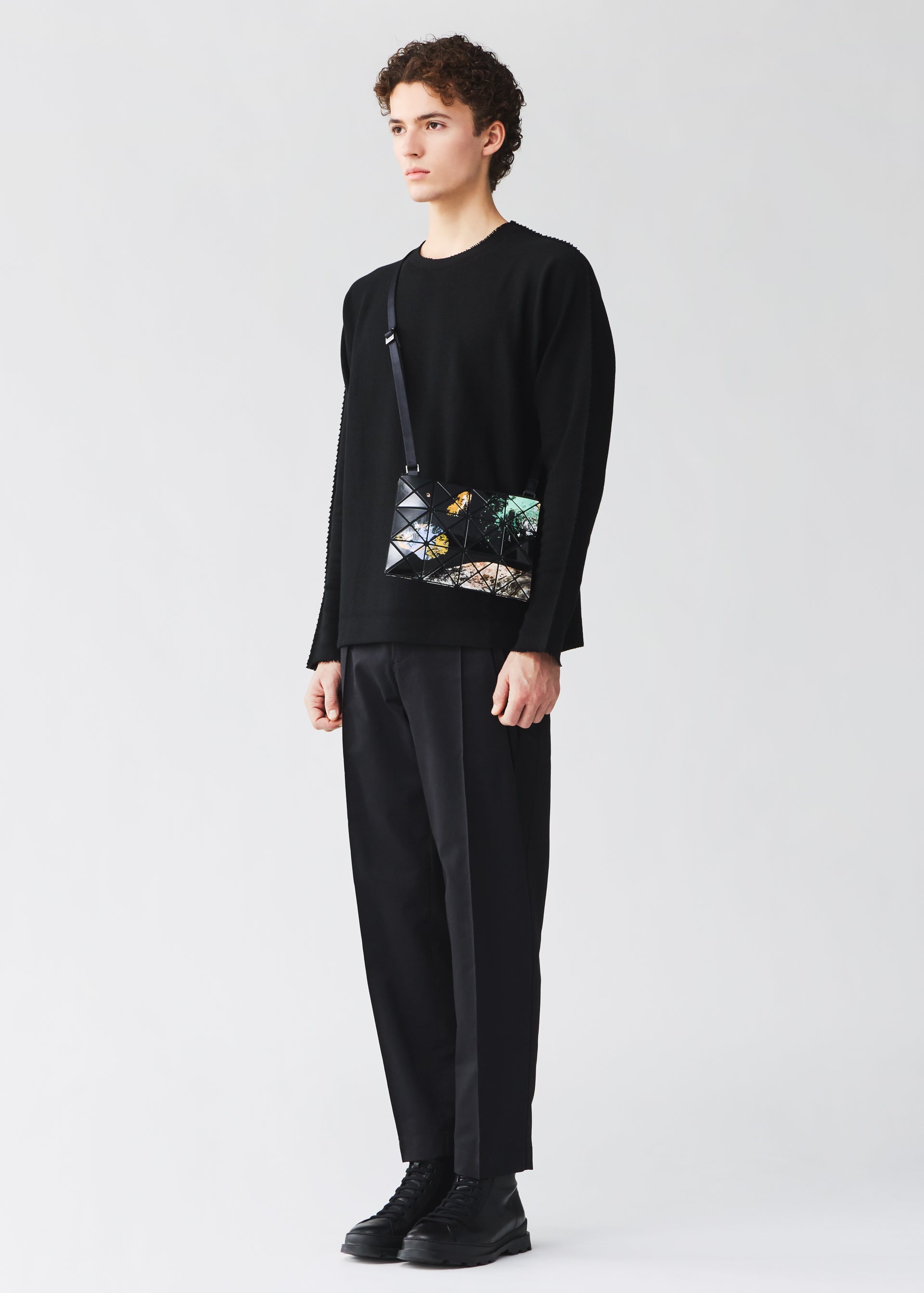Zoomed view A person stands against a plain background, wearing a black sweater, black pants, black shoes, and carrying the BAO BAO ISSEY MIYAKE COSMIC shoulder bag, which features a geometric design with colorful accents and an adjustable strap for added comfort and versatility.