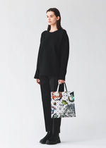 A person wearing a loose black sweater, black pants, and black shoes stands holding the COSMIC tote bag by BAO BAO ISSEY MIYAKE, its glossy enamel material and colorful abstract geometric patterns standing out against the plain light background.