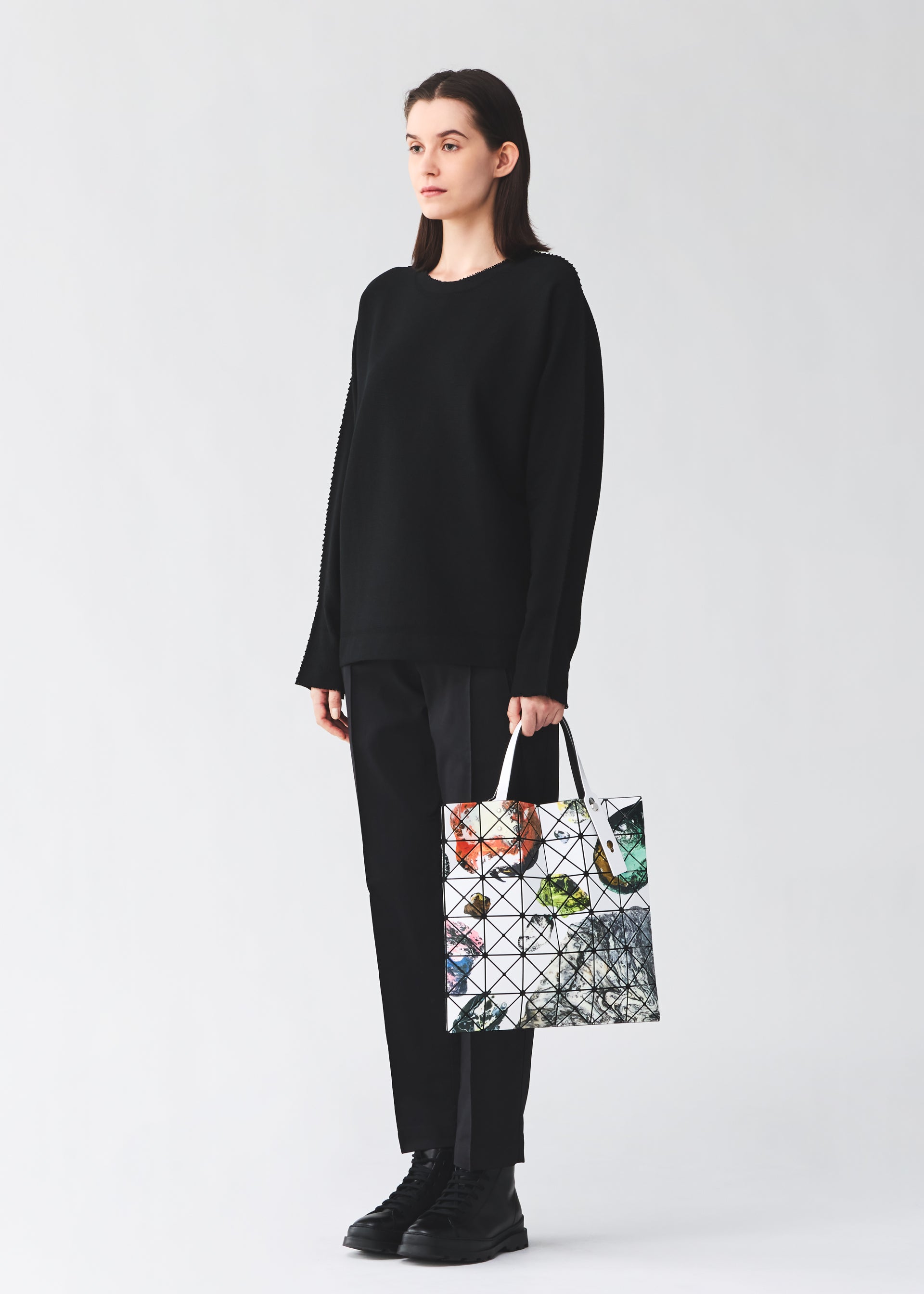 Zoomed view A person wearing a loose black sweater, black pants, and black shoes stands holding the COSMIC tote bag by BAO BAO ISSEY MIYAKE, its glossy enamel material and colorful abstract geometric patterns standing out against the plain light background.