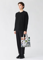A person dressed in black—sweater, pants, and shoes—stands against a plain light background, holding the COSMIC tote bag from BAO BAO ISSEY MIYAKE. The spacious enamel bag features a geometric pattern with colorful accents and fits A4-sized documents.