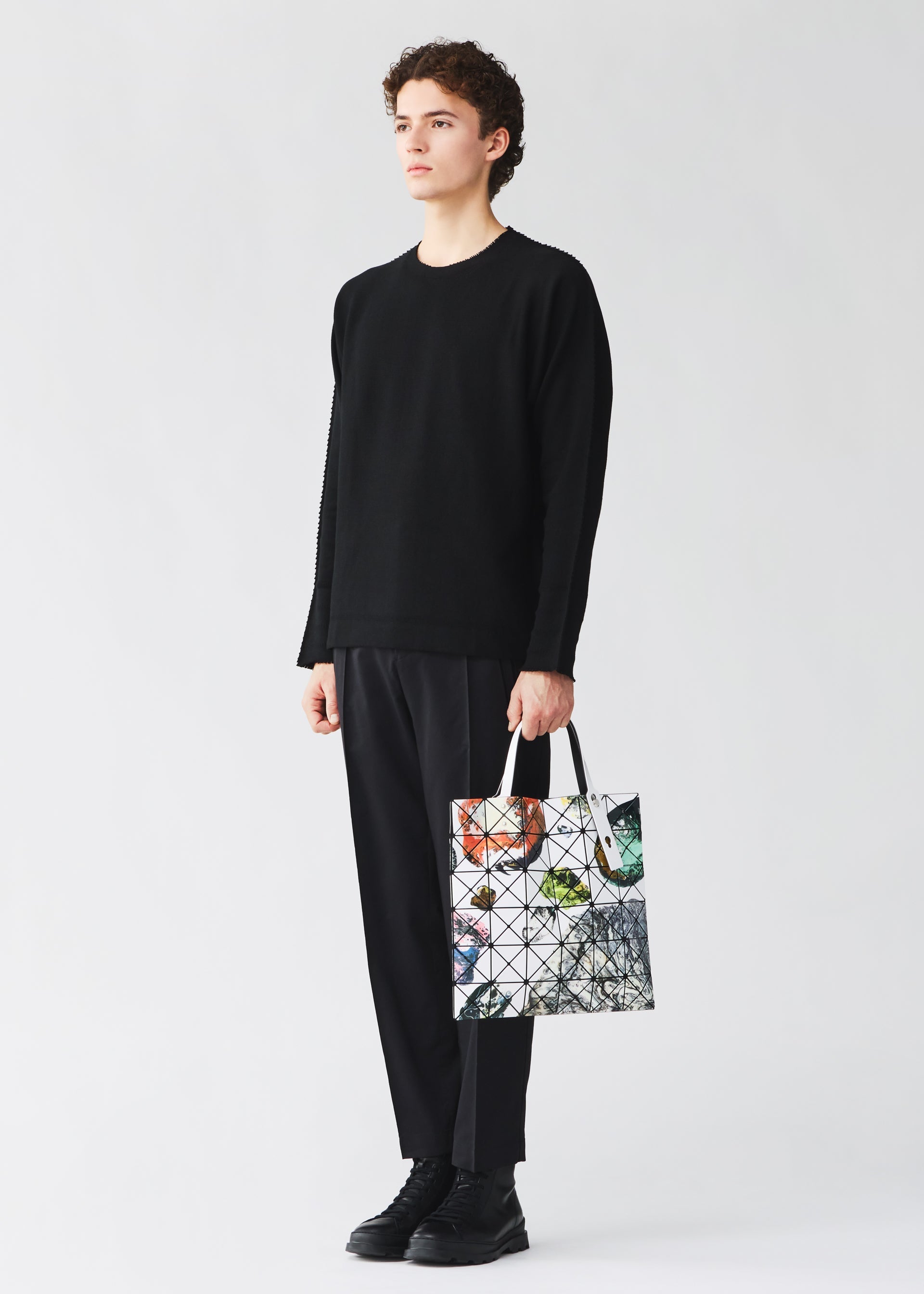 Zoomed view A person dressed in black—sweater, pants, and shoes—stands against a plain light background, holding the COSMIC tote bag from BAO BAO ISSEY MIYAKE. The spacious enamel bag features a geometric pattern with colorful accents and fits A4-sized documents.