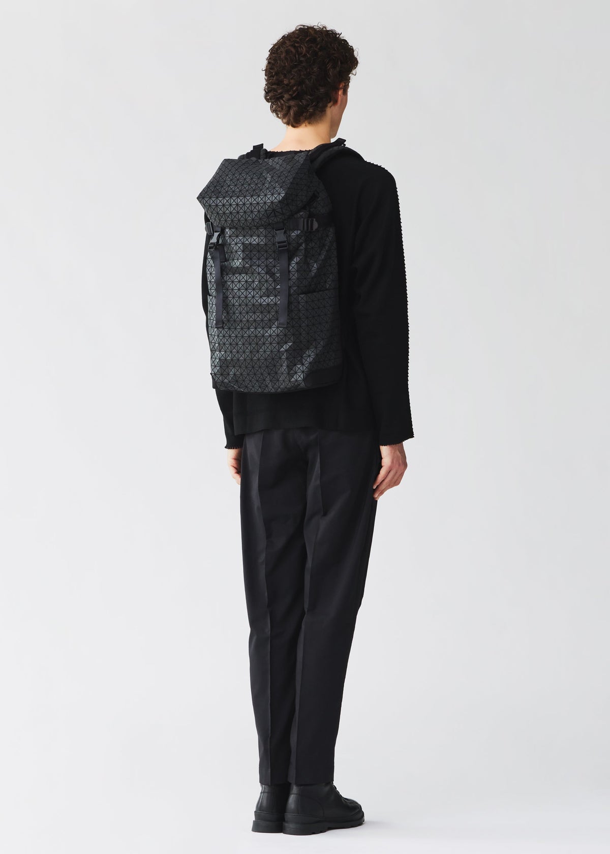 A person with short curly hair, in black clothing and shoes, stands facing away wearing a BAO BAO ISSEY MIYAKE HIKER backpack with a laptop pocket against a plain light background. view 7 of 8