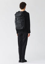 A person with short curly hair, in black clothing and shoes, stands facing away wearing a BAO BAO ISSEY MIYAKE HIKER backpack with a laptop pocket against a plain light background.