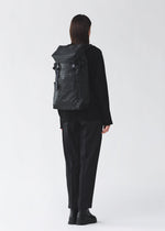 A person with short dark hair in black clothing and boots stands facing away, wearing a BAO BAO ISSEY MIYAKE HIKER backpack with a laptop pocket, against a plain light gray background.
