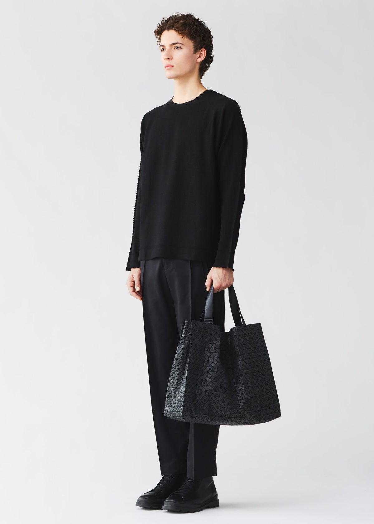 A person with short curly hair stands against a plain light background, dressed in black and holding the BAO BAO ISSEY MIYAKE CART tote bag with adjustable handles in their left hand. view 5 of 6