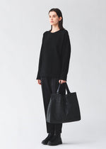 A person with straight brown hair wears a loose black sweater, black pants, and shoes, standing against a light background while holding the BAO BAO ISSEY MIYAKE CART tote bag with adjustable handles.