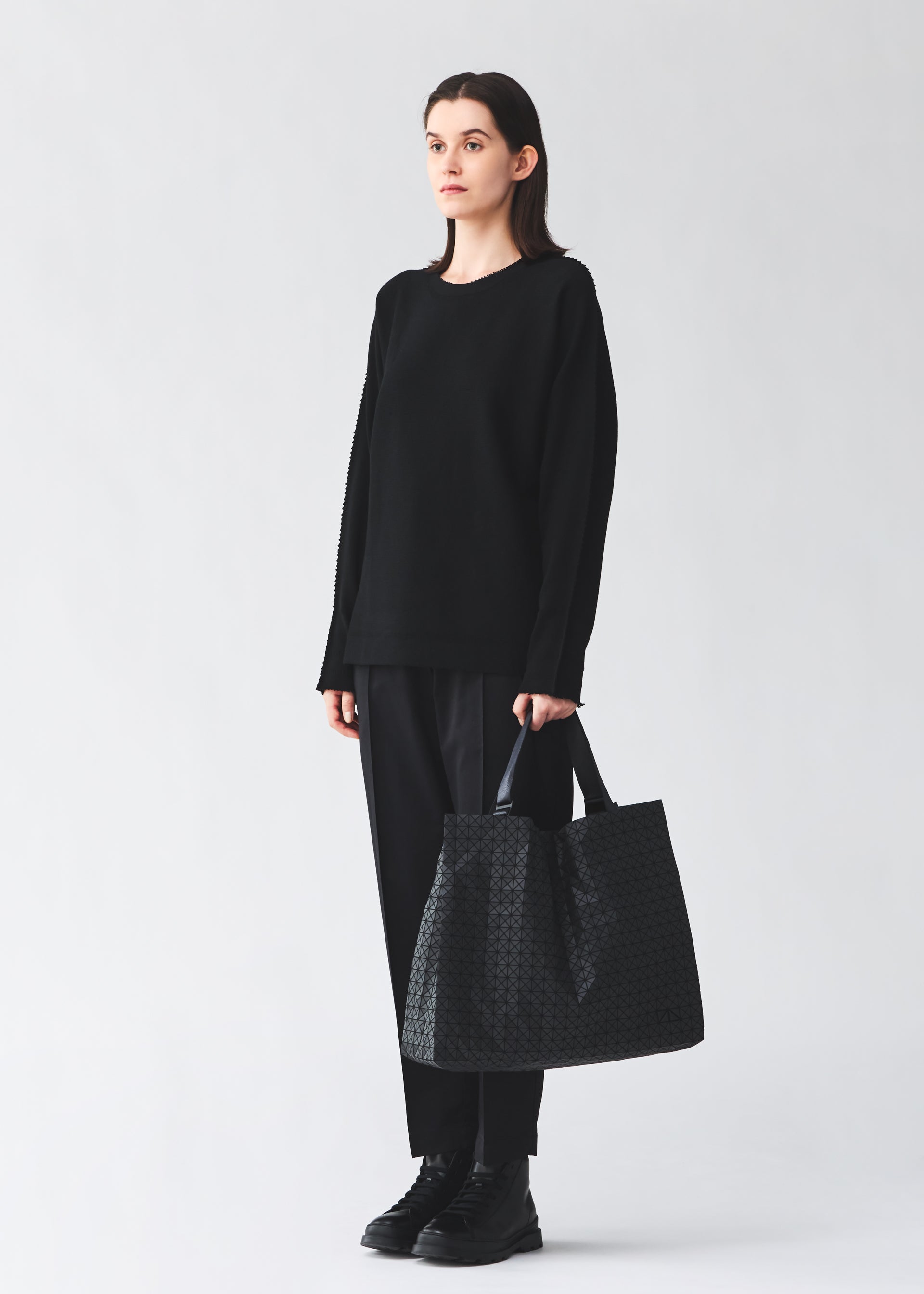 Zoomed view A person with straight brown hair wears a loose black sweater, black pants, and shoes, standing against a light background while holding the BAO BAO ISSEY MIYAKE CART tote bag with adjustable handles.