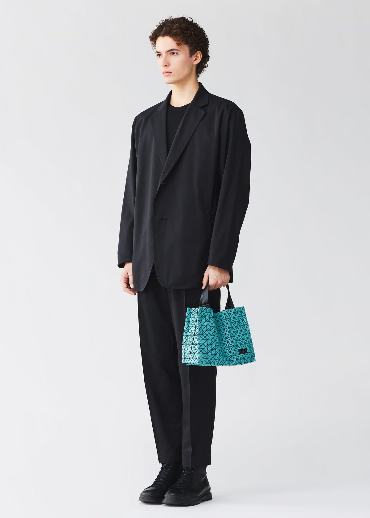 Zoomed view A person in a loose black suit holds the CART MINI by BAO BAO ISSEY MIYAKE, a small turquoise patterned tote, standing against a plain white background with a neutral expression.