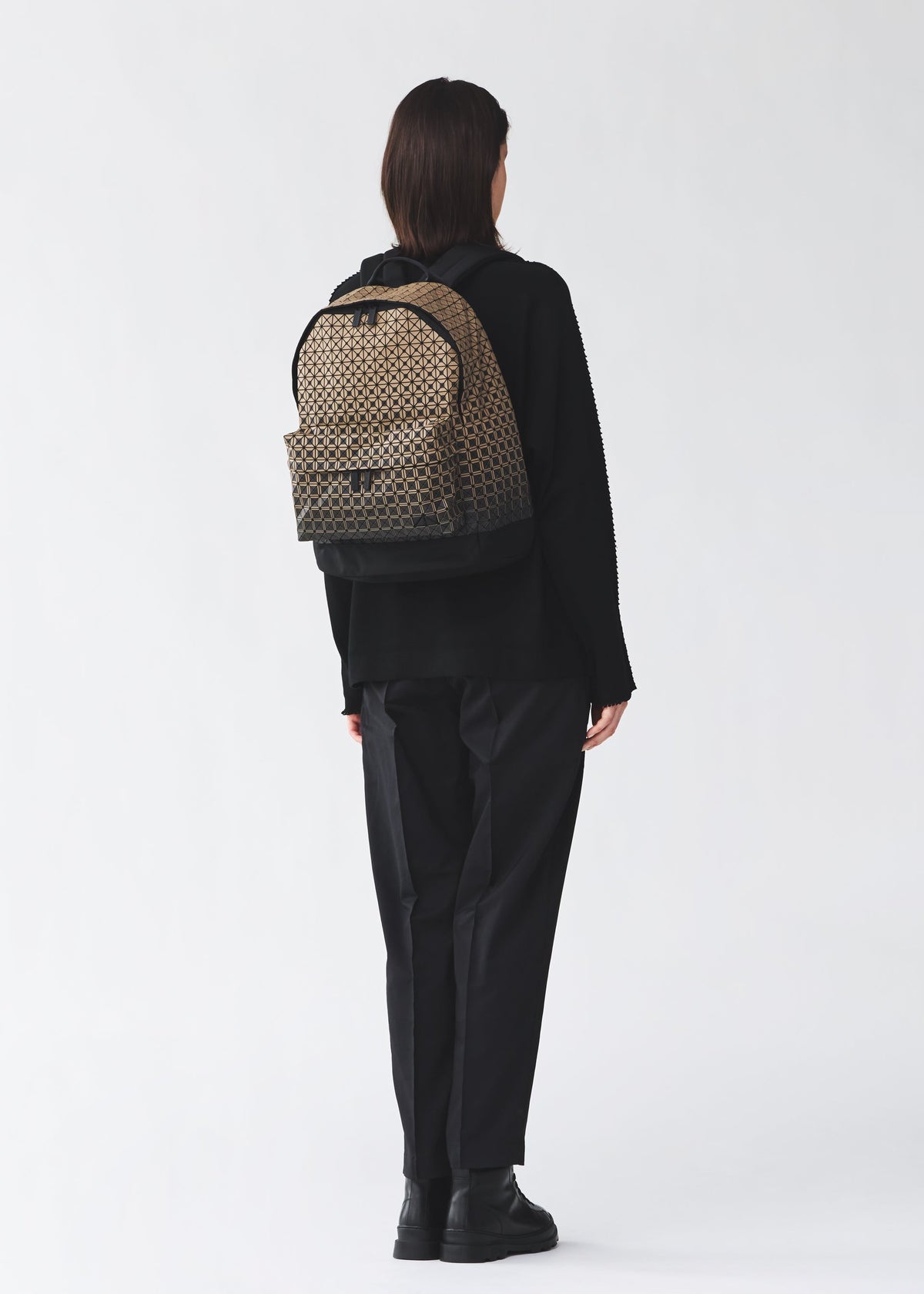A person stands facing away, dressed in black clothing and black boots, carrying the BAO BAO ISSEY MIYAKE DAYPACK GRADUAL—a beige and black patterned backpack showcasing a striking graphic gradation—against a plain light background. view 7 of 7