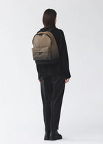 A person stands facing away, dressed in black clothing and black boots, carrying the BAO BAO ISSEY MIYAKE DAYPACK GRADUAL—a beige and black patterned backpack showcasing a striking graphic gradation—against a plain light background.