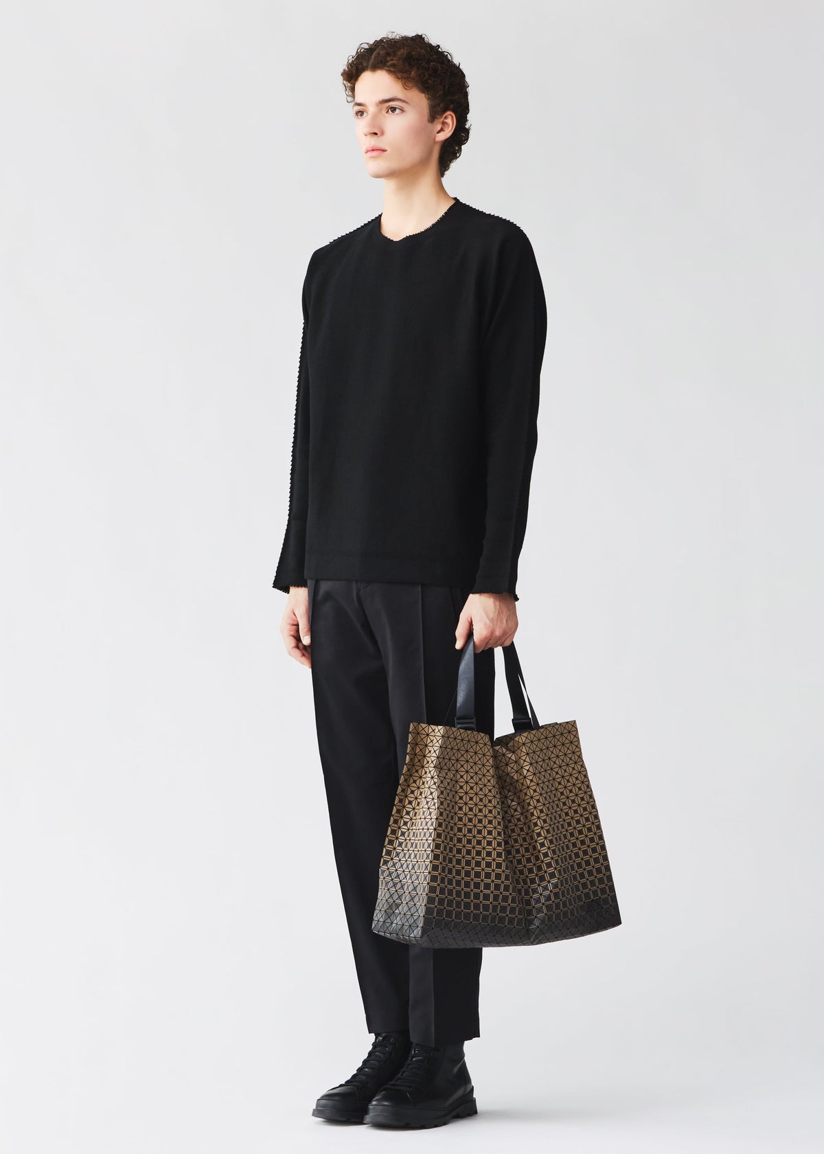 A person with curly hair, dressed in a black long-sleeve shirt, black pants, and black shoes, stands against a plain light background holding the CART GRADUAL—a large brown patterned tote bag by BAO BAO ISSEY MIYAKE featuring adjustable handles and designed to perfectly carry A4-sized documents. view 7 of 8