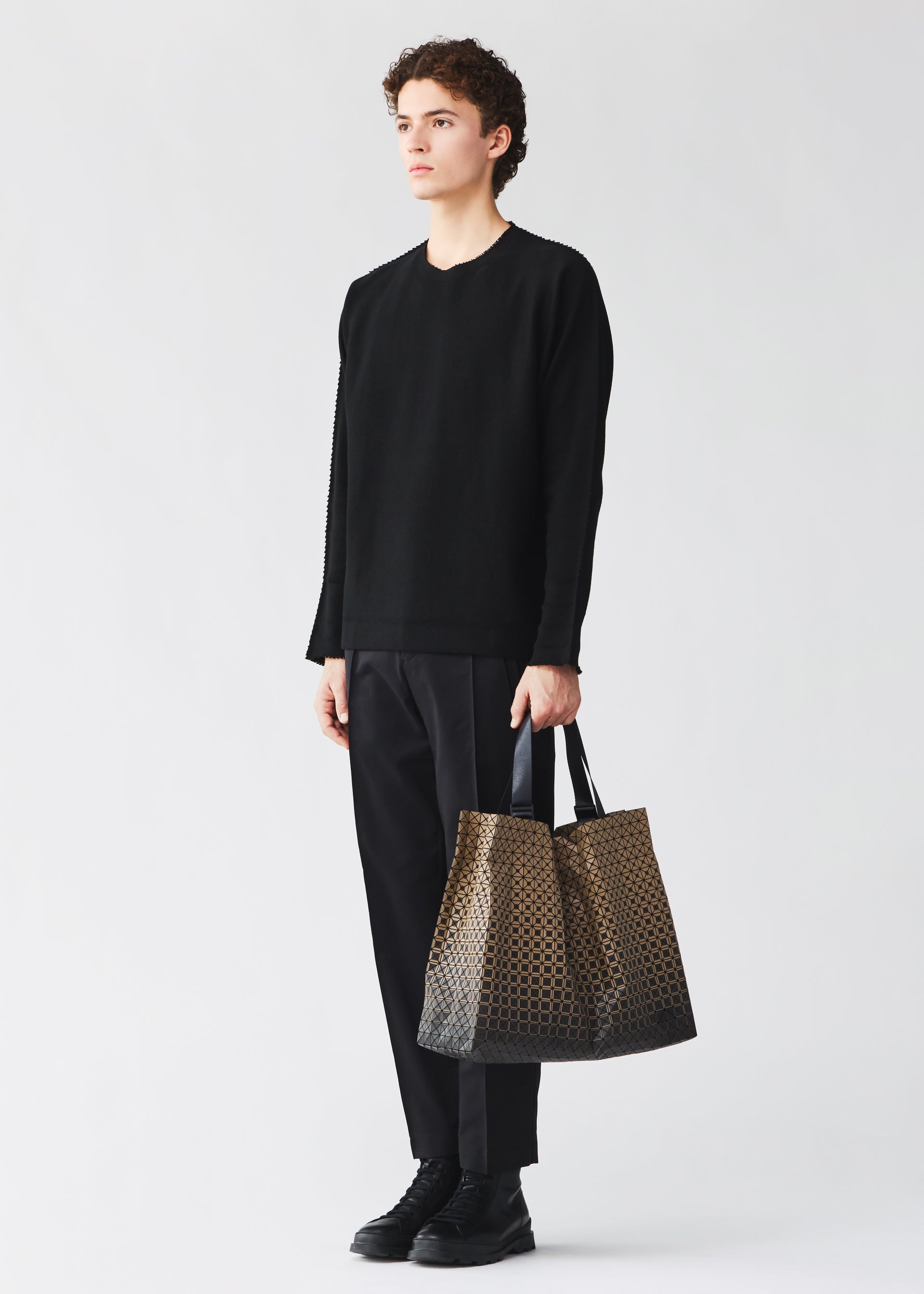 Zoomed view A person with curly hair, dressed in a black long-sleeve shirt, black pants, and black shoes, stands against a plain light background holding the CART GRADUAL—a large brown patterned tote bag by BAO BAO ISSEY MIYAKE featuring adjustable handles and designed to perfectly carry A4-sized documents.
