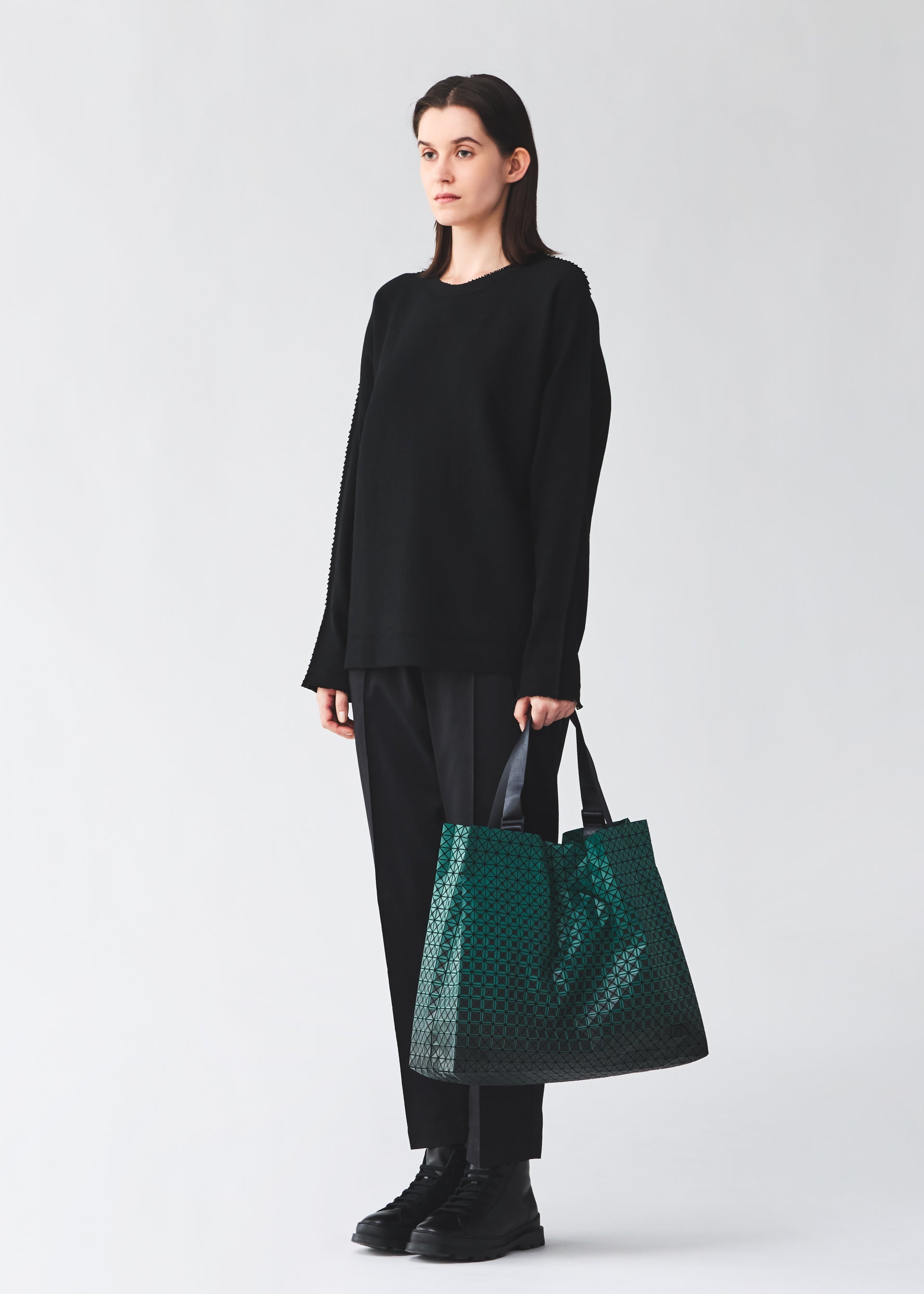 Zoomed view A person with long dark hair stands facing forward, dressed in a loose black sweater, black pants, and black boots. They are holding the CART GRADUAL—an A4-sized green patterned tote bag with adjustable handles by BAO BAO ISSEY MIYAKE—ideal for carrying documents. The background is plain and light-colored.