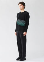 A person wearing a black long-sleeve top, loose black pants, and black shoes stands against a plain light background, holding the BAO BAO ISSEY MIYAKE SADDLE BAG GRADUAL—a green patterned box-shaped shoulder bag.