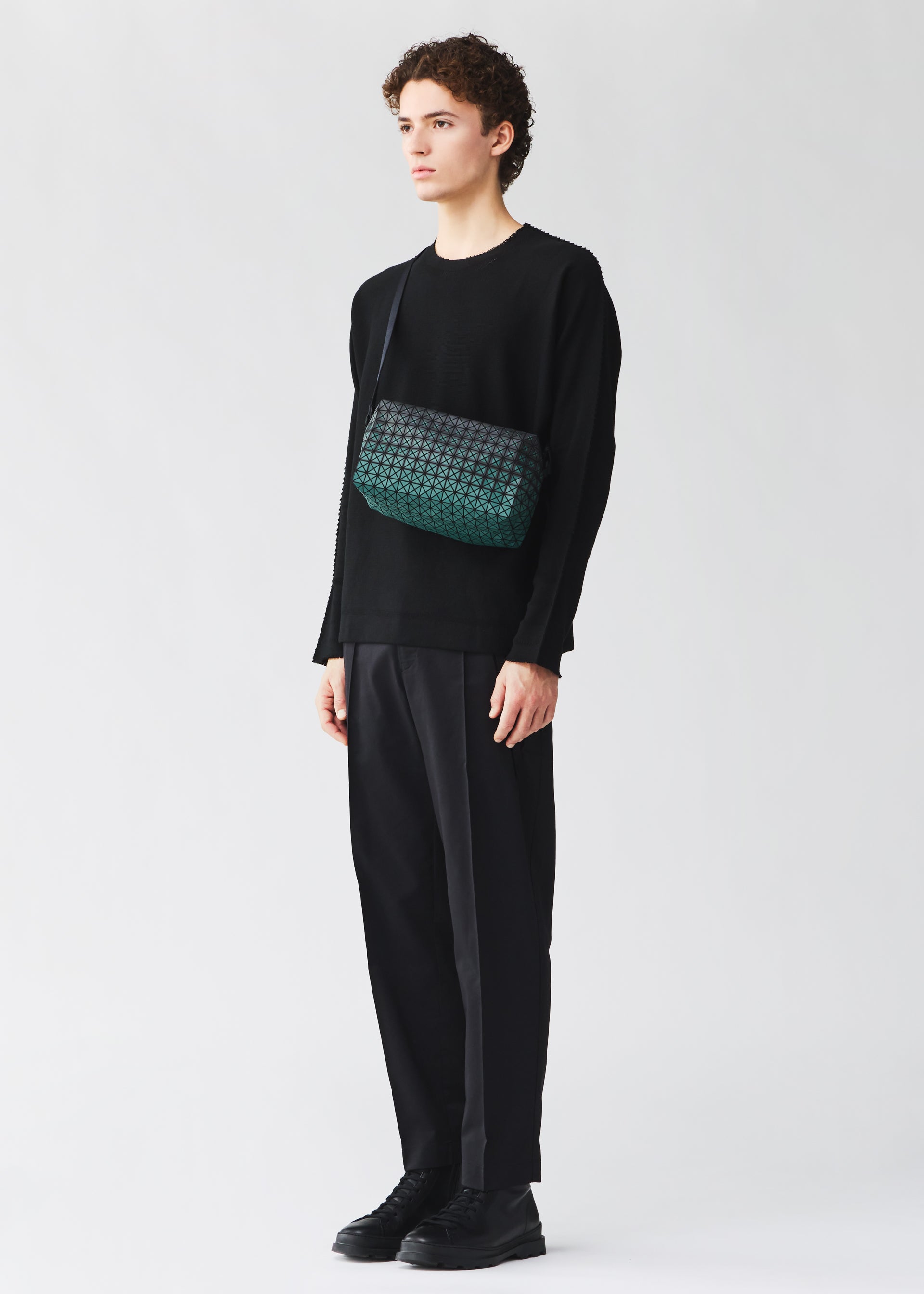 Zoomed view A person wearing a black long-sleeve top, loose black pants, and black shoes stands against a plain light background, holding the BAO BAO ISSEY MIYAKE SADDLE BAG GRADUAL—a green patterned box-shaped shoulder bag.