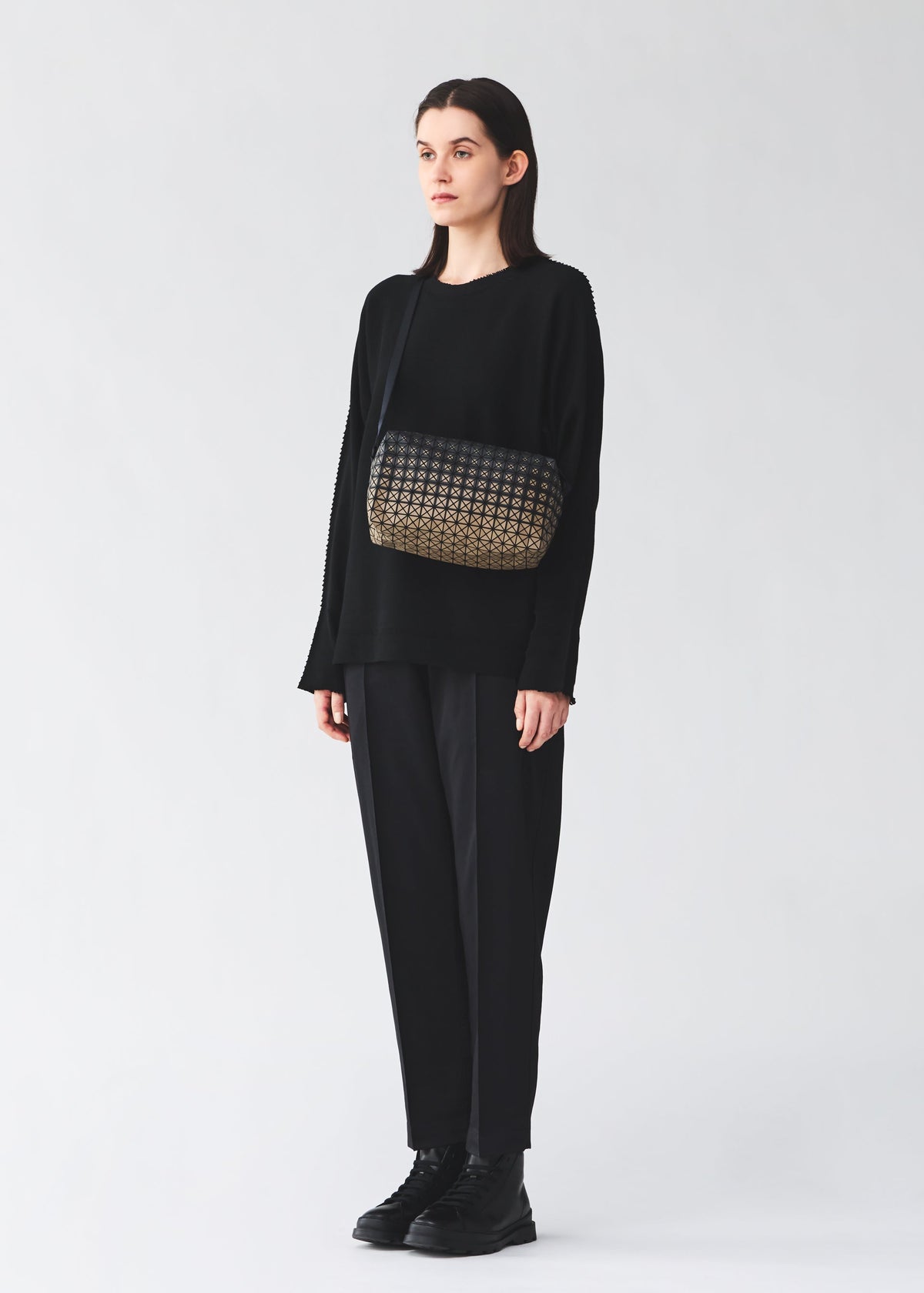 A person wearing a loose black outfit and black shoes stands against a light background, carrying the BAO BAO ISSEY MIYAKE SADDLE BAG GRADUAL from the KURO series—a box-shaped shoulder bag featuring a patterned black and gold design. view 8 of 8