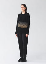 A person wearing a loose black outfit and black shoes stands against a light background, carrying the BAO BAO ISSEY MIYAKE SADDLE BAG GRADUAL from the KURO series—a box-shaped shoulder bag featuring a patterned black and gold design.