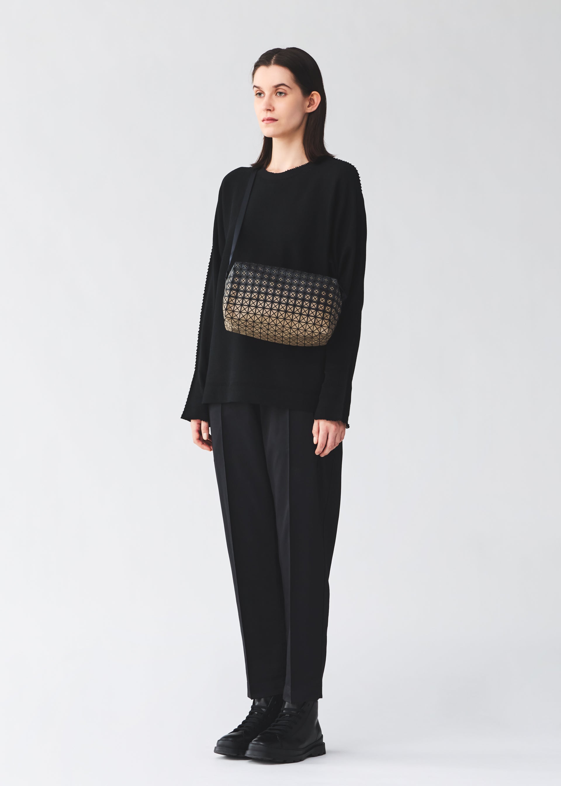 Zoomed view A person wearing a loose black outfit and black shoes stands against a light background, carrying the BAO BAO ISSEY MIYAKE SADDLE BAG GRADUAL from the KURO series—a box-shaped shoulder bag featuring a patterned black and gold design.