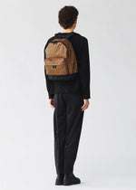 A person with short curly hair, dressed in all-black attire, stands facing away against a plain light background, wearing the BAO BAO ISSEY MIYAKE DAYPACK REFLECTOR— a three-dimensional backpack with brown patterns.