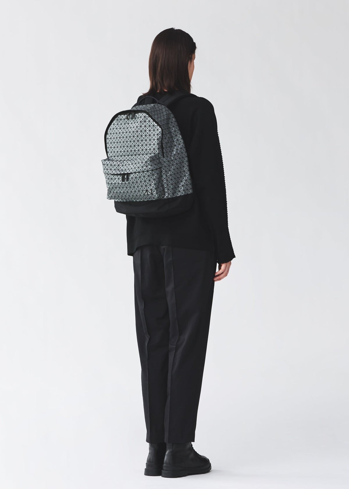 A person with short dark hair, dressed in loose black clothing and boots, stands facing away from the camera wearing the light gray DAYPACK REFLECTOR by BAO BAO ISSEY MIYAKE. The background is plain and light-colored. view 6 of 7