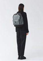 A person with short dark hair, dressed in loose black clothing and boots, stands facing away from the camera wearing the light gray DAYPACK REFLECTOR by BAO BAO ISSEY MIYAKE. The background is plain and light-colored.