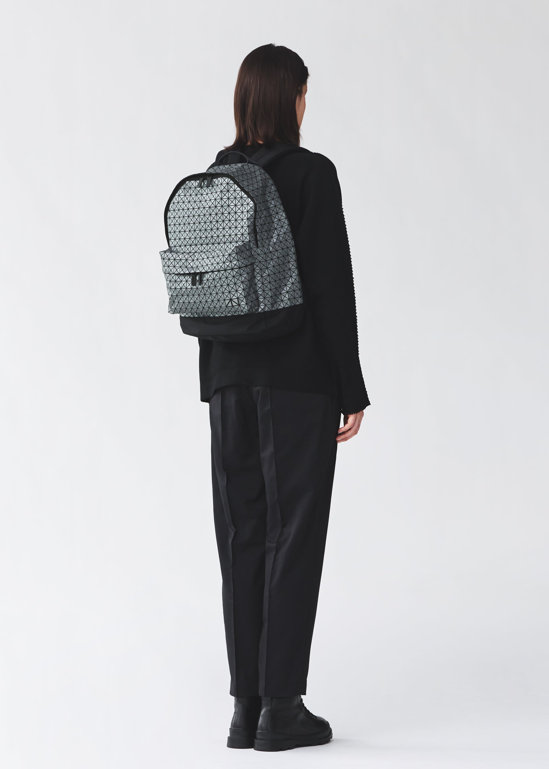 Zoomed view A person with short dark hair, dressed in loose black clothing and boots, stands facing away from the camera wearing the light gray DAYPACK REFLECTOR by BAO BAO ISSEY MIYAKE. The background is plain and light-colored.