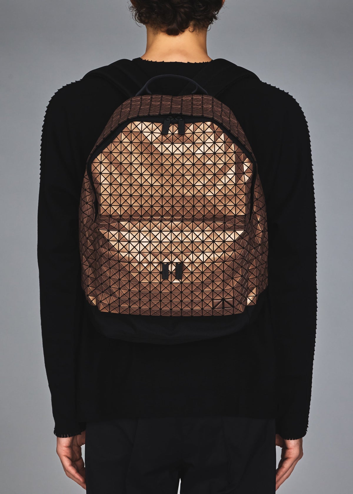 A person in black attire is seen from behind carrying the BAO BAO ISSEY MIYAKE DAYPACK REFLECTOR, a three-dimensional backpack featuring a brown and black geometric triangular grid design, against a plain gray background. view 7 of 7
